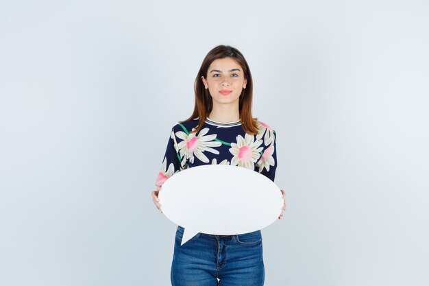 Chica joven sosteniendo bocadillo en blusa floral, jeans y mirando complacido, vista frontal.