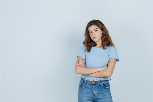 Chica joven sosteniendo los brazos cruzados en camiseta, jeans y mirando serio, vista frontal.