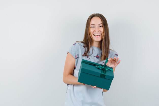 Chica joven sosteniendo la caja actual en camiseta y mirando feliz. vista frontal.