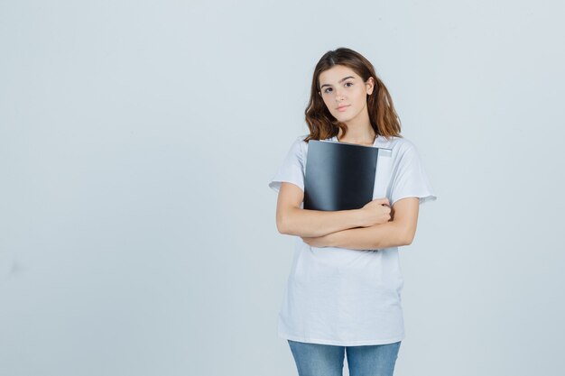 Chica joven sosteniendo una carpeta en camiseta blanca y mirando sensible, vista frontal.