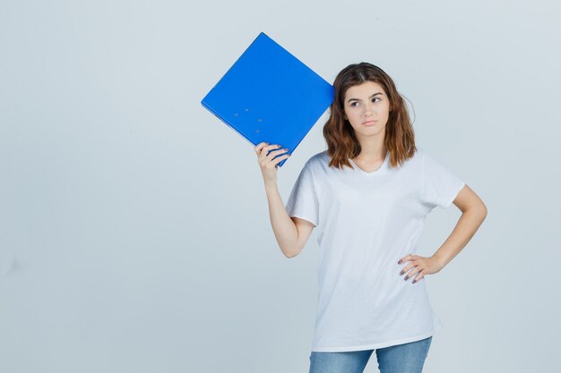 Chica joven sosteniendo la carpeta, manteniendo la mano en la cintura en camiseta blanca y mirando vacilante, vista frontal.