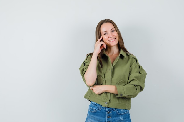 Chica joven sosteniendo el dedo en la mejilla en chaqueta, pantalones cortos y mirando alegre, vista frontal.