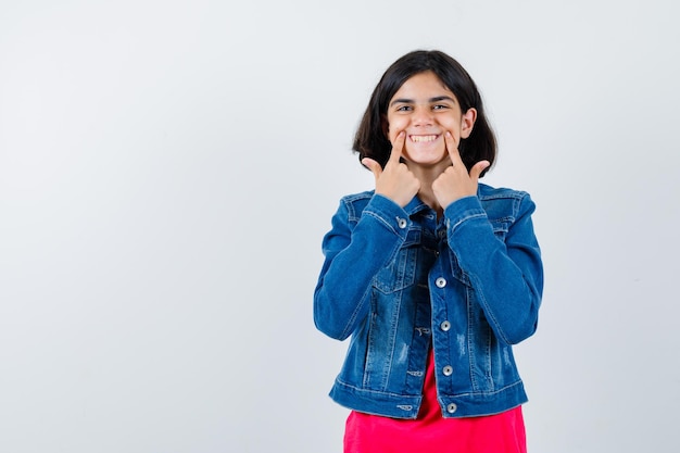 Chica joven sosteniendo los dedos índices cerca de la boca, forzando una sonrisa en camiseta roja y chaqueta de jean y luciendo feliz, vista frontal.
