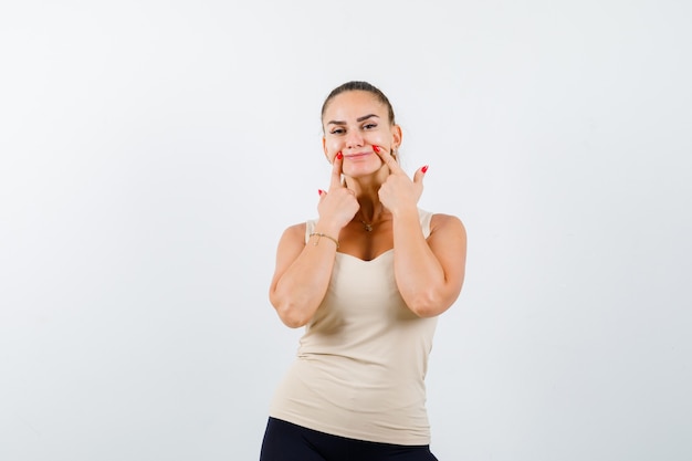 Chica joven sosteniendo los dedos índices cerca de la boca, forzando una sonrisa en la parte superior beige, pantalón negro y luciendo seria. vista frontal.