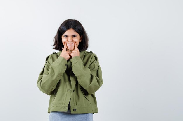 Chica joven sosteniendo los dedos índices cerca de la boca, forzando una sonrisa en suéter gris, chaqueta caqui, pantalón de mezclilla y mirando serio, vista frontal.