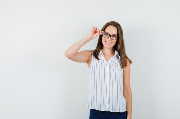 Chica joven sosteniendo los dedos en los vidrios en camiseta, jeans y mirando jovial, vista frontal.