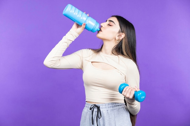 Chica joven sosteniendo mancuernas y bebiendo agua Joven chica en forma de pie sobre fondo púrpura Foto de alta calidad