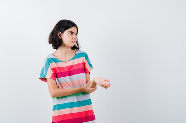 Chica joven sosteniendo la mano en el antebrazo, mirando a otro lado en una camiseta a rayas de colores y mirando seriamente, vista frontal.