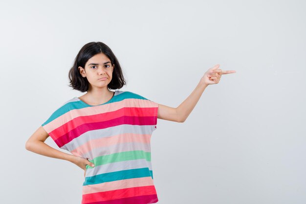 Chica joven sosteniendo la mano en la cintura mientras apunta a la derecha en una camiseta a rayas de colores y mirando serio, vista frontal.