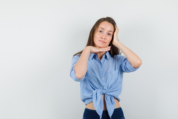 Chica joven sosteniendo la mano debajo de la barbilla en camisa azul, pantalones y luciendo linda