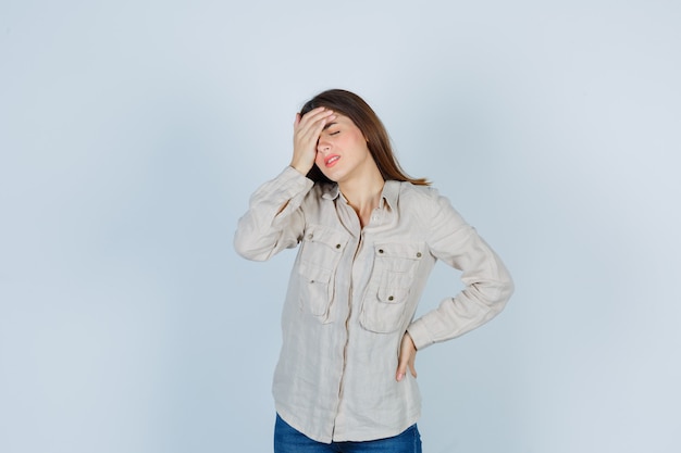 Foto gratuita chica joven sosteniendo una mano en la frente, otra mano en la cadera en camisa beige, jeans y mirando acosado. vista frontal.