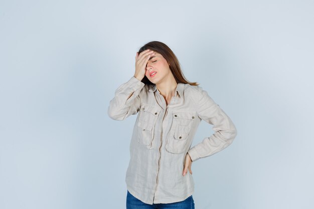 Chica joven sosteniendo una mano en la frente, otra mano en la cadera en camisa beige, jeans y mirando acosado. vista frontal.