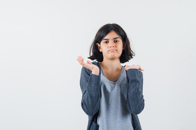 Chica joven sosteniendo una mano en el hombro, estirando la otra mano en una camiseta gris claro y una sudadera con capucha gris oscuro con cremallera y luciendo linda.