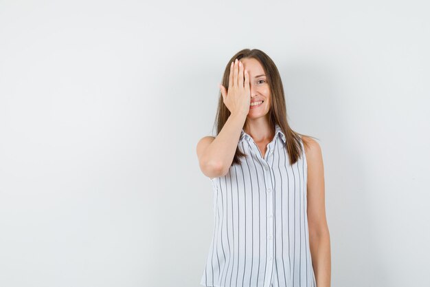 Chica joven sosteniendo la mano en el ojo en camiseta y mirando positivo, vista frontal.