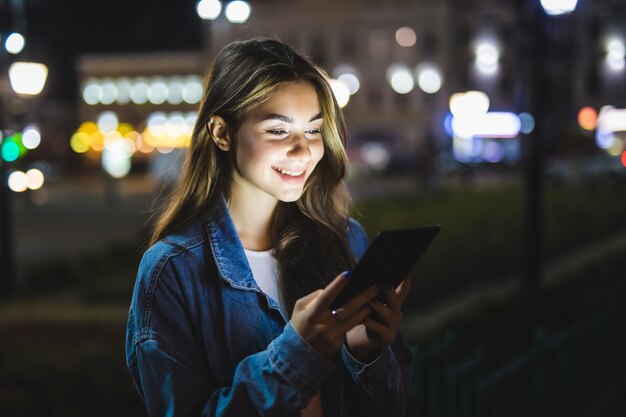 Chica joven sosteniendo en las manos en la tableta de pantalla en blanco por la noche.