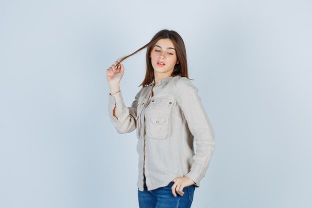 Chica joven sosteniendo un mechón de cabello, con la mano en la cadera mientras posa en camisa beige, jeans y luce atractivo. vista frontal.