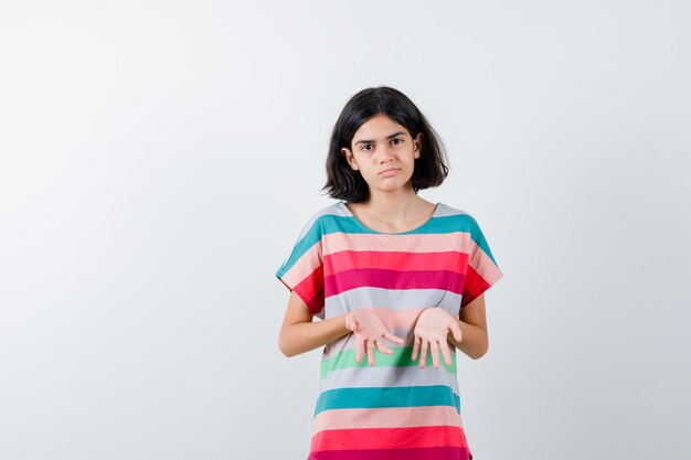 Chica joven sosteniendo las palmas en camiseta a rayas de colores y mirando serio, vista frontal.