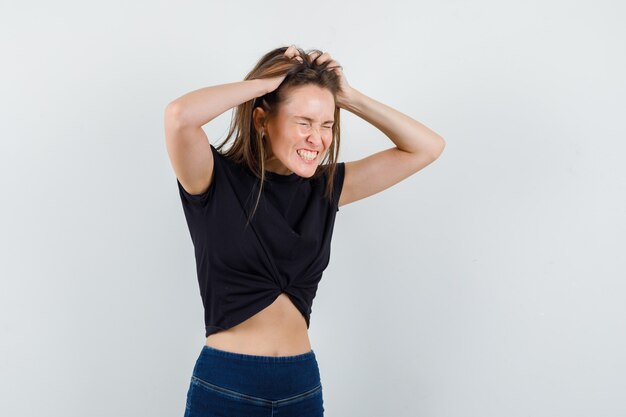 Chica joven sosteniendo el pelo con las manos en blusa negra, pantalones y mirando dichoso