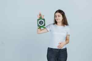 Foto gratuita chica joven sosteniendo el reloj mientras mantiene la mano en el estómago en camiseta, jeans y mirando confiado, vista frontal.