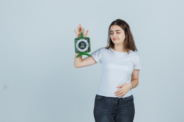 Chica joven sosteniendo el reloj mientras mantiene la mano en el estómago en camiseta, jeans y mirando confiado, vista frontal.