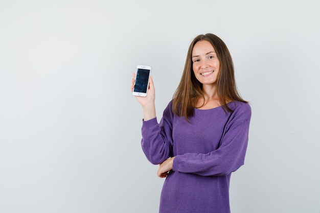 Chica joven sosteniendo teléfono móvil en camisa violeta y mirando alegre, vista frontal.