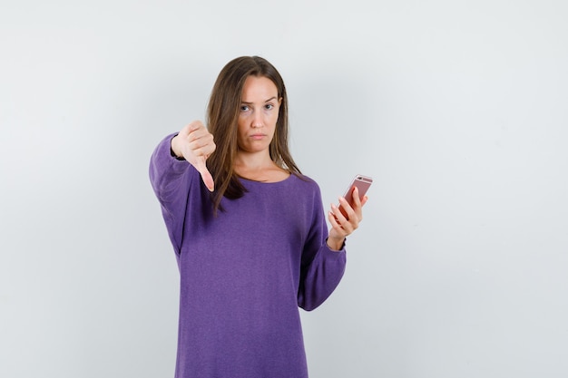 Chica joven sosteniendo el teléfono móvil con el pulgar hacia abajo en camisa violeta y mirando decepcionado. vista frontal.