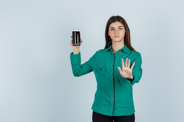 Chica joven sosteniendo un vaso de líquido negro, mostrando la señal de pare en blusa verde, pantalón negro y mirando serio. vista frontal.