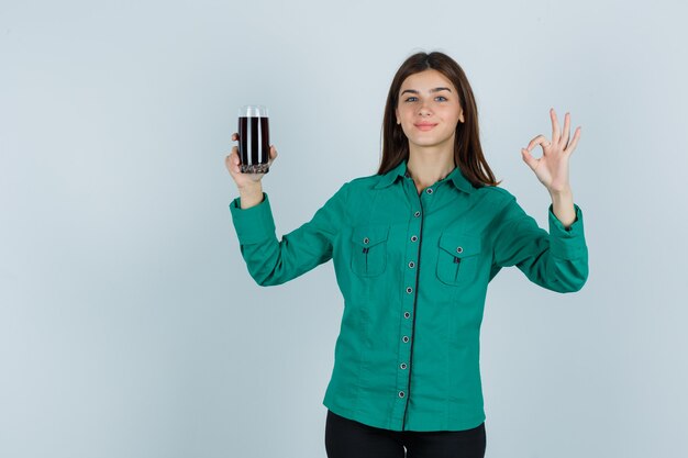 Chica joven sosteniendo un vaso de líquido negro, mostrando el signo de ok en blusa verde, pantalón negro y luciendo feliz. vista frontal.