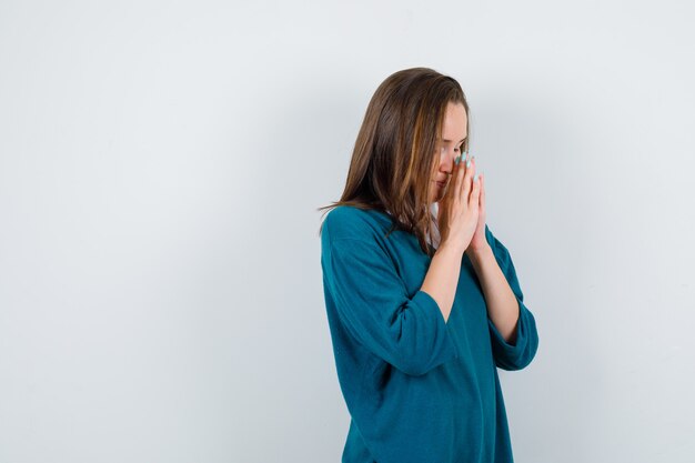 Chica joven en suéter con cuello en v, camisa que muestra el gesto de namaste, de pie de lado y mirando agradecido.