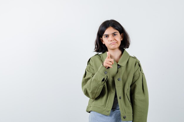 Chica joven en suéter gris, chaqueta caqui, pantalón de jean apuntando y mirando serio, vista frontal.