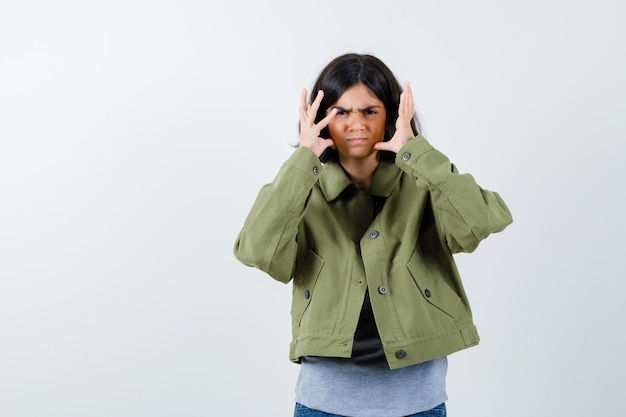 Chica joven en suéter gris, chaqueta caqui, pantalón de mezclilla cogidos de la mano cerca de la cabeza, haciendo muecas y mirando acosado, vista frontal.