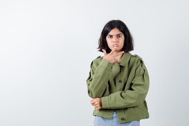 Chica joven en suéter gris, chaqueta caqui, pantalón de mezclilla estirando la mano hacia la cámara y mirando serio, vista frontal.