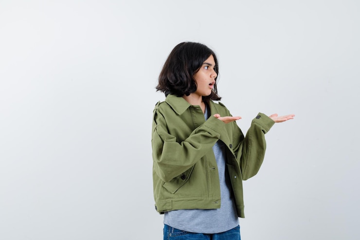 Foto gratuito chica joven en suéter gris, chaqueta caqui, pantalón de mezclilla estirando las manos como sosteniendo algo imaginario y mirando sorprendido, vista frontal.