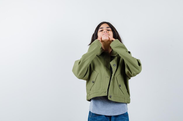 Chica joven en suéter gris, chaqueta caqui, pantalón de mezclilla sosteniendo los dedos índices cerca de la boca, forzando una sonrisa y luciendo linda vista frontal.