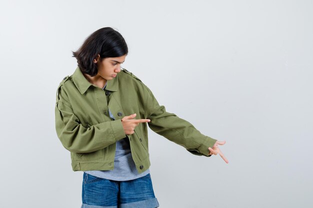 Chica joven en suéter gris, chaqueta de color caqui, pantalón de mezclilla apuntando hacia abajo y hacia la derecha con los dedos índices y mirando enfocado, vista frontal.