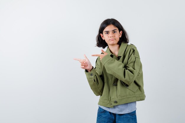 Chica joven en suéter gris, chaqueta de color caqui, pantalón de mezclilla apuntando hacia la izquierda con los dedos índices y mirando serio, vista frontal.