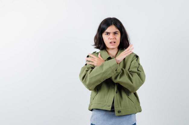 Chica joven en suéter gris, chaqueta de color caqui, pantalón de mezclilla con dos brazos cruzados, gesticulando sin señal y mirando serio, vista frontal.