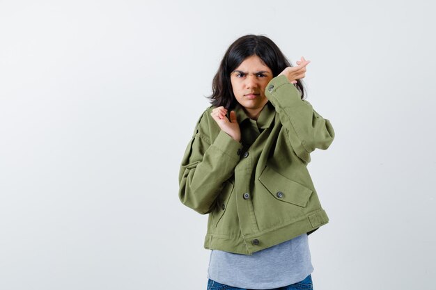 Chica joven en suéter gris, chaqueta de color caqui, pantalón de mezclilla estirando las manos de manera interrogante y mirando perplejo, vista frontal.