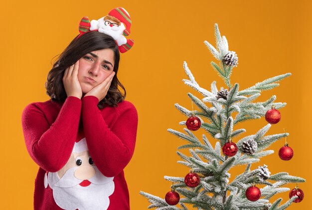 Chica joven en suéter de Navidad con diadema divertida mirando a la cámara con expresión triste en la cara de pie junto a un árbol de Navidad sobre fondo naranja