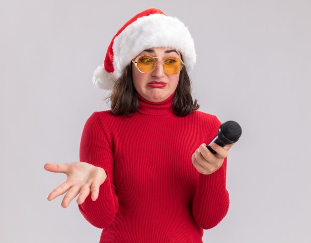 Chica joven en suéter rojo y gorro de Papá Noel con gafas sosteniendo micrófono mirándolo con expresión confusa con el brazo extendido sobre fondo blanco.