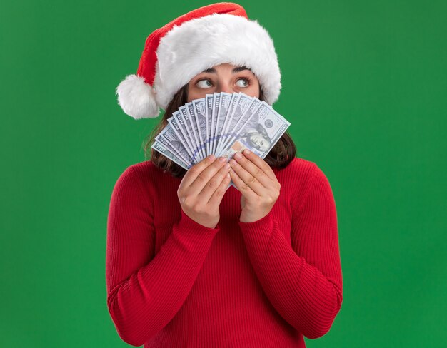 Chica joven en suéter rojo y gorro de Papá Noel que cubre la cara con dinero mirando a un lado preocupado sosteniendo efectivo sobre fondo verde