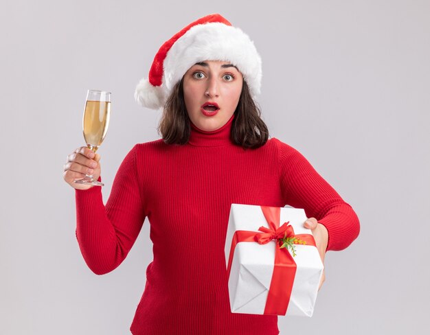 Chica joven en suéter rojo y gorro de Papá Noel sosteniendo una copa de champán y un presente mirando a la cámara sorprendido de pie sobre fondo blanco.