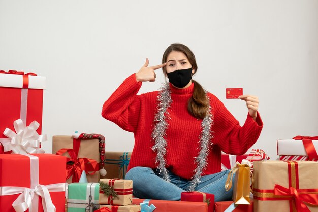 Chica joven con suéter rojo y máscara negra poniendo la pistola de dedo en la sien sentada alrededor de regalos en blanco