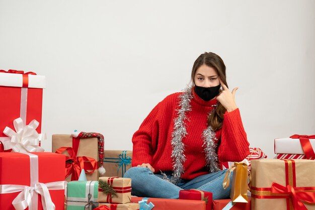 Chica joven con suéter rojo y máscara negra poniendo la pistola de dedo en la sien sentada alrededor de regalos en blanco