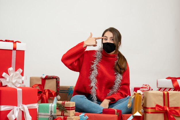 Foto gratuita chica joven con suéter rojo y máscara negra poniendo la pistola de dedo en la sien sentada alrededor de regalos en blanco