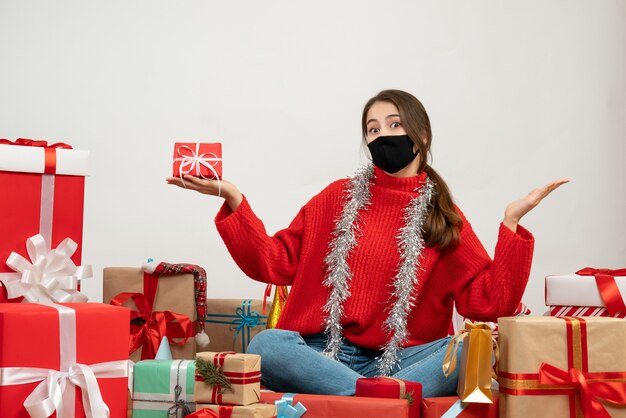 Chica joven con suéter rojo y máscara negra sosteniendo giftbox sentados alrededor presenta en blanco