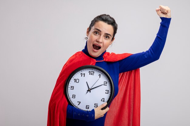 Chica joven superhéroe alegre sosteniendo un reloj de pared y mostrando un gesto de sí aislado sobre fondo blanco.