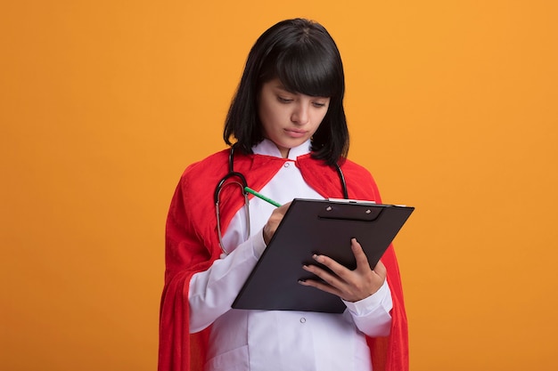 Chica joven superhéroe confiada con estetoscopio con bata médica y manto escribiendo algo en el portapapeles