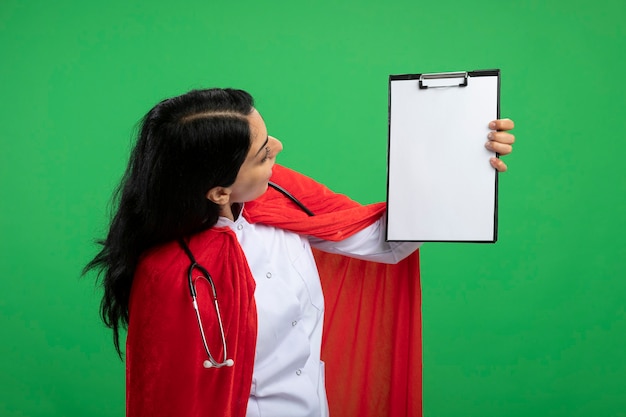 Chica joven superhéroe sorprendida con bata médica con estetoscopio levantando y mirando el portapapeles aislado en verde