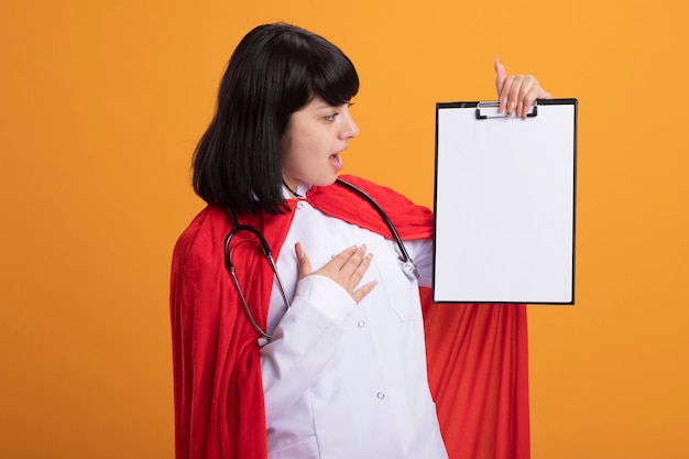 Foto gratuita chica joven superhéroe sorprendida con estetoscopio con bata médica y manto sosteniendo y mirando el portapapeles poniendo la mano en el corazón aislado en naranja